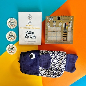 May include: A blue and white fish-shaped fabric pouch with a black eye, next to a gold circuit board with the text "Mon Khum" and "Soundscape Machine". There are three round stickers with the text "Mon Khum" in a yellow and orange background.