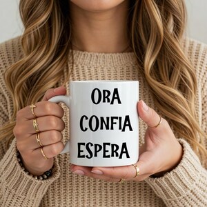 Puede incluir: Taza de cerámica blanca con las palabras "ORA CONFIA ESPERA" en negro. La taza es sostenida por una persona. La taza tiene un asa estándar y una superficie lisa. La persona lleva un suéter beige y anillos dorados.