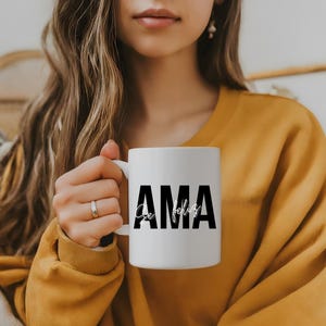 Puede incluir: Taza de cerámica blanca con la palabra "AMA" en letras negras en negrita y "Se feliz" en fuente cursiva. La taza es sostenida por una persona que lleva un jersey amarillo.