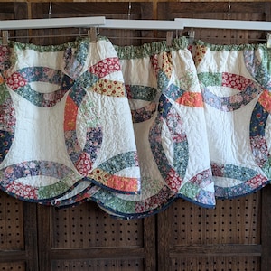 May include: Three skirts made from quilted fabric featuring a double wedding ring pattern. The skirts have an elastic waistband, a scalloped hemline, and are constructed from white fabric with multicolored floral print rings in red, green, and blue.