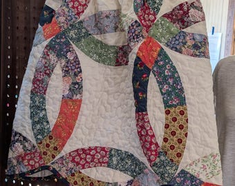 May include: A quilted skirt featuring a double wedding ring pattern with various floral fabric patches in red, green, blue, and yellow. The skirt has a cream-colored quilted background, a green floral elastic waistband, and a scalloped hemline.
