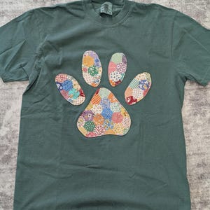 May include: A green t-shirt featuring a colorful patchwork paw print design. The paw print is made of various fabric scraps in different colors and patterns. The shirt is a comfortable, casual style.