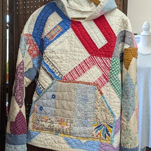 May include: A long-sleeve hooded sweatshirt constructed from various quilted fabric patches. The garment features a front kangaroo pocket and a mix of patterns including floral, plaid, and geometric prints in red, blue, yellow, and cream.