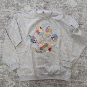May include: A light gray sweatshirt with a colorful patchwork paw print design. The paw print is made of various fabric patterns and colors, including red, blue, yellow, and green. The sweatshirt has a crew neck and long sleeves.