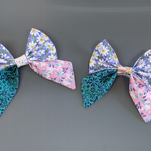 May include: Two fabric hair bows. Each bow features a combination of patterns: a teal floral print, a pink floral print, and a blue daisy print. The bows are tied in the center with a small fabric piece.