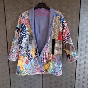 May include: A patchwork jacket with a variety of colorful fabric patterns. The jacket has a light blue lining and collar, and is hanging on a black hanger.