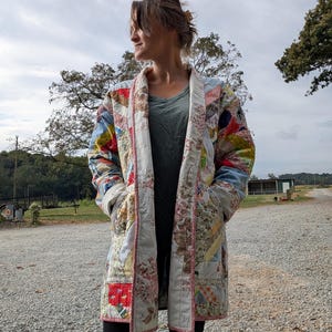May include: A long-sleeved coat constructed from a patchwork quilt featuring a variety of floral and geometric patterns in shades of blue, red, yellow, and white. The coat has a wide collar, two front pockets, and pink trim along the edges.
