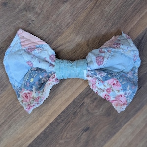 May include: A fabric bow featuring a patchwork design of light blue, pink, and white floral patterns. The edges of the bow have a frayed, raw appearance, and the center is cinched with a solid light blue fabric band, creating a textured look.