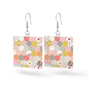 May include: A pair of square earrings with a patchwork design. The earrings feature a variety of hexagonal patterns in shades of pink, yellow, orange, and gray, with floral and checkered details. The earrings hang from silver-colored hooks.