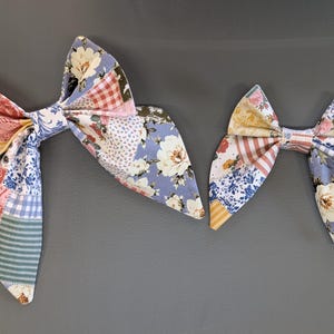 May include: Two fabric hair bows with a patchwork design. The bows feature a mix of floral, gingham, and striped patterns in shades of red, blue, pink, and green. The bows are likely handmade and suitable for various occasions.