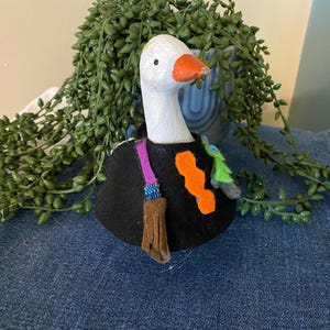 May include: A white goose figurine with an orange beak wearing a black felt cape. The cape is decorated with a purple strap, a brown fringed pouch, a bright orange wavy felt shape, and a small green felt accent attached to the front.