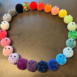 May include: A collection of twenty-four small, round, crocheted plush spheres arranged in a circle. Each sphere features a soft, textured yarn surface in a different color, including red, orange, yellow, green, blue, purple, pink, and black.