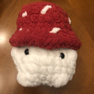 May include: A white crocheted mushroom plush toy with a red and white crocheted cap. The mushroom has black button eyes.