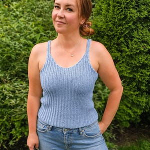 Può includere: Una canotta azzurra senza maniche con una trama a maglia a coste verticali, uno scollo rotondo e spalline sottili. Il capo è aderente e arriva fino alla vita, abbinato a un classico paio di jeans blu con chiusura a bottone e cerniera.
