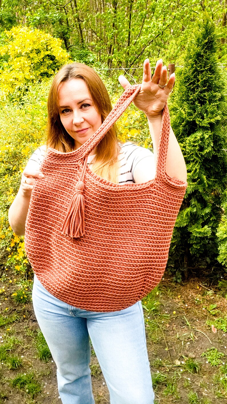 May include: A woman is holding a crocheted tote bag with a tassel. The bag is a light brown color and has a simple, textured design. The woman is wearing a striped shirt and blue jeans.