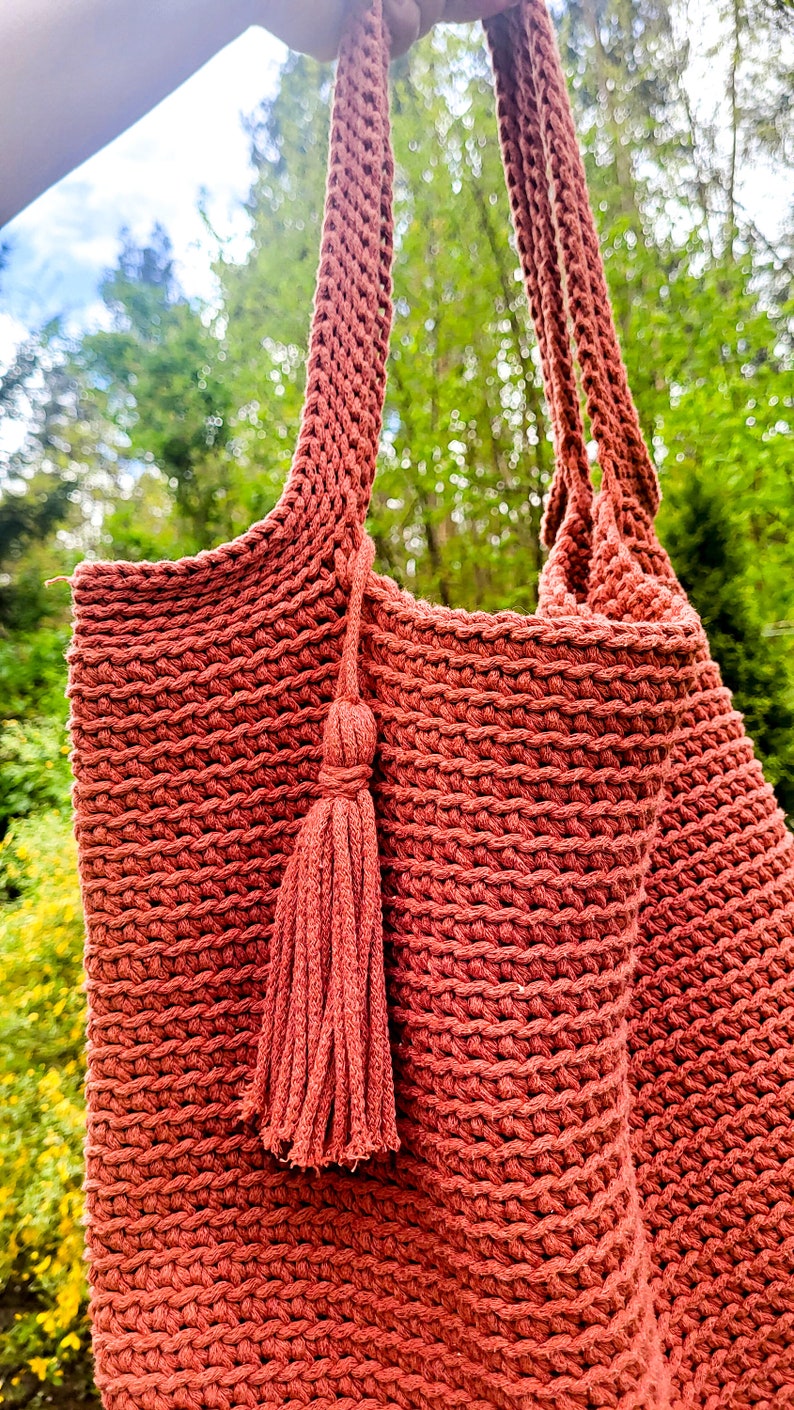 May include: A terracotta-colored crocheted tote bag with a tassel. The bag is made with a thick, textured yarn and has a simple, classic design.