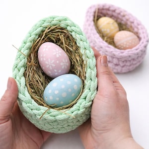 May include: A light green crocheted egg-shaped basket filled with straw and two decorated Easter eggs. One egg is pink with white floral designs, and the other is light blue with white polka dots. A second basket with two eggs is in the background.