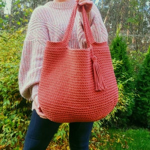 May include: A large, crocheted tote bag in a terracotta color with a matching tassel. The bag is made with a simple stitch pattern and has a wide, open top.