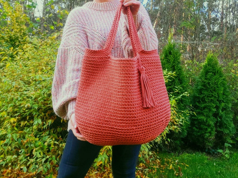May include: A large, crocheted tote bag in a terracotta color with a matching tassel. The bag is made with a simple stitch pattern and has a wide, open top.