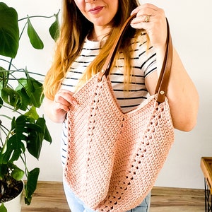 Può includere: Una borsa tote rosa in crochet con manici in pelle marrone. La borsa è tenuta da una persona che indossa una camicia a righe e jeans blu.