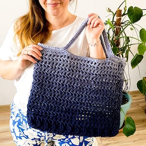 Puede incluir: Una mujer sostiene una bolsa de mano de crochet con un degradado de hilo azul y morado. La bolsa tiene una parte superior ancha y abierta y un asa resistente. La mujer lleva una camisa blanca y pantalones de flores azules y blancos.