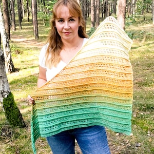 Op de afbeelding: Een vrouw in een wit shirt en blauwe jeans houdt een gehaakte sjaal vast met een gradiënt van groen, geel en wit.
