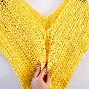 Puede incluir: Un top de crochet amarillo con escote en V. El top está colgado en una percha de madera sobre un fondo blanco.