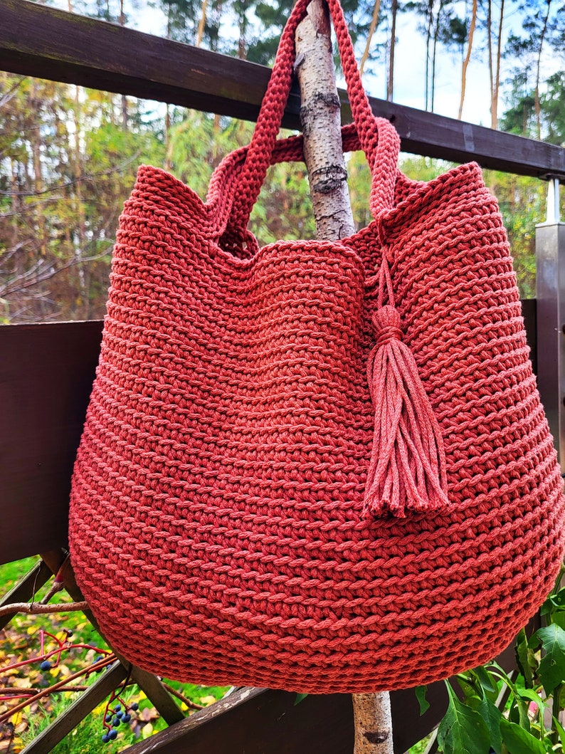May include: A large, red crocheted tote bag with a tassel hanging from the side. The bag is hanging from a branch in a wooded area.