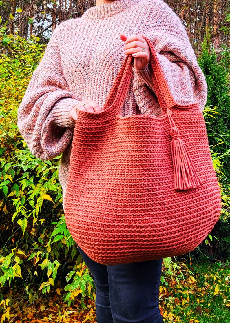 May include: A large, crocheted tote bag with a tassel. The bag is a deep coral color and is made with a thick yarn. The bag is being held by a person wearing a pink sweater and jeans.