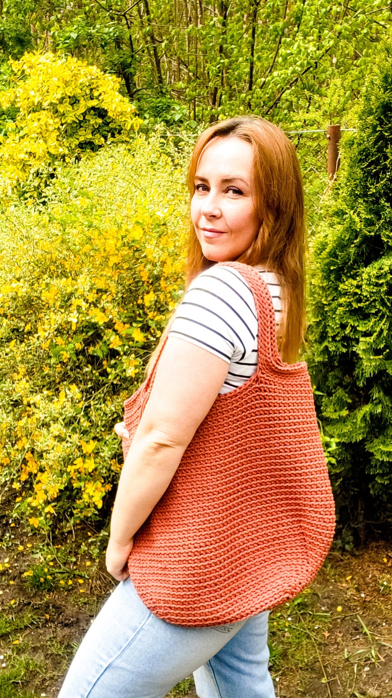 May include: A woman wearing a striped shirt and blue jeans is carrying a large, crocheted tote bag in a terracotta color.