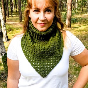 Puede incluir: Una mujer lleva una bufanda triangular de ganchillo verde con detalles blancos. Está de pie en un entorno boscoso.