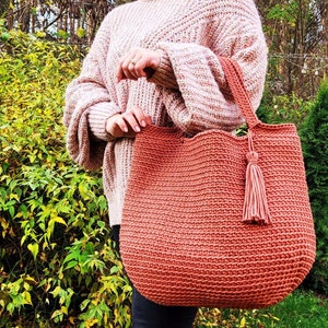 May include: A large, crocheted tote bag with a tassel. The bag is a deep orange color and is made with a thick yarn. The bag is being held by a person wearing a pink sweater.
