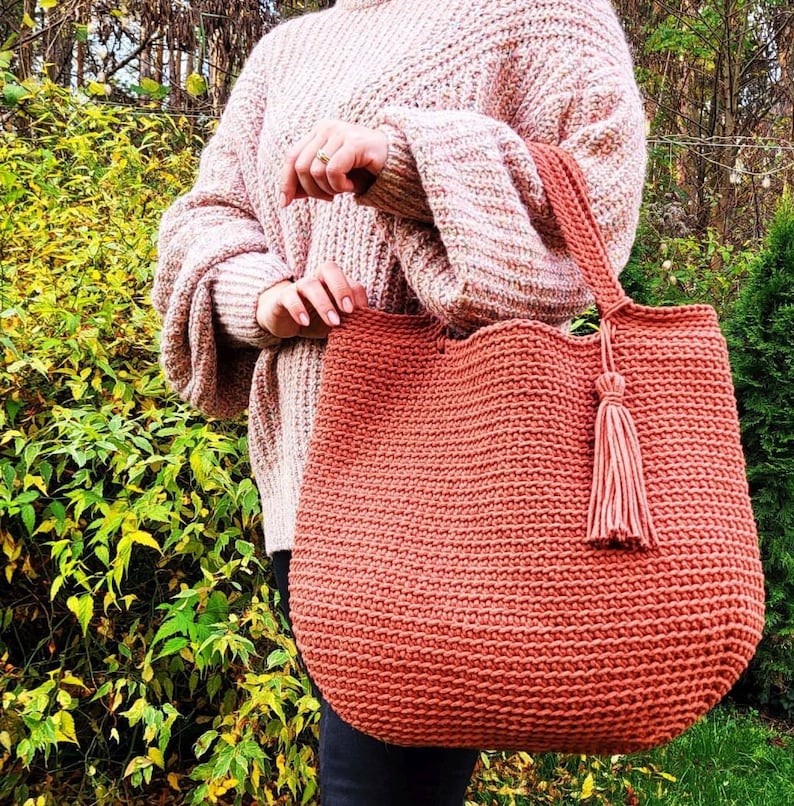 May include: A large, crocheted tote bag with a tassel. The bag is a deep orange color and is made with a thick yarn. The bag is being held by a person wearing a pink sweater.