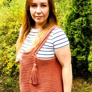 May include: A woman is wearing a striped shirt and blue jeans. She is holding a large, crocheted tote bag with a tassel. The bag is a terracotta color.