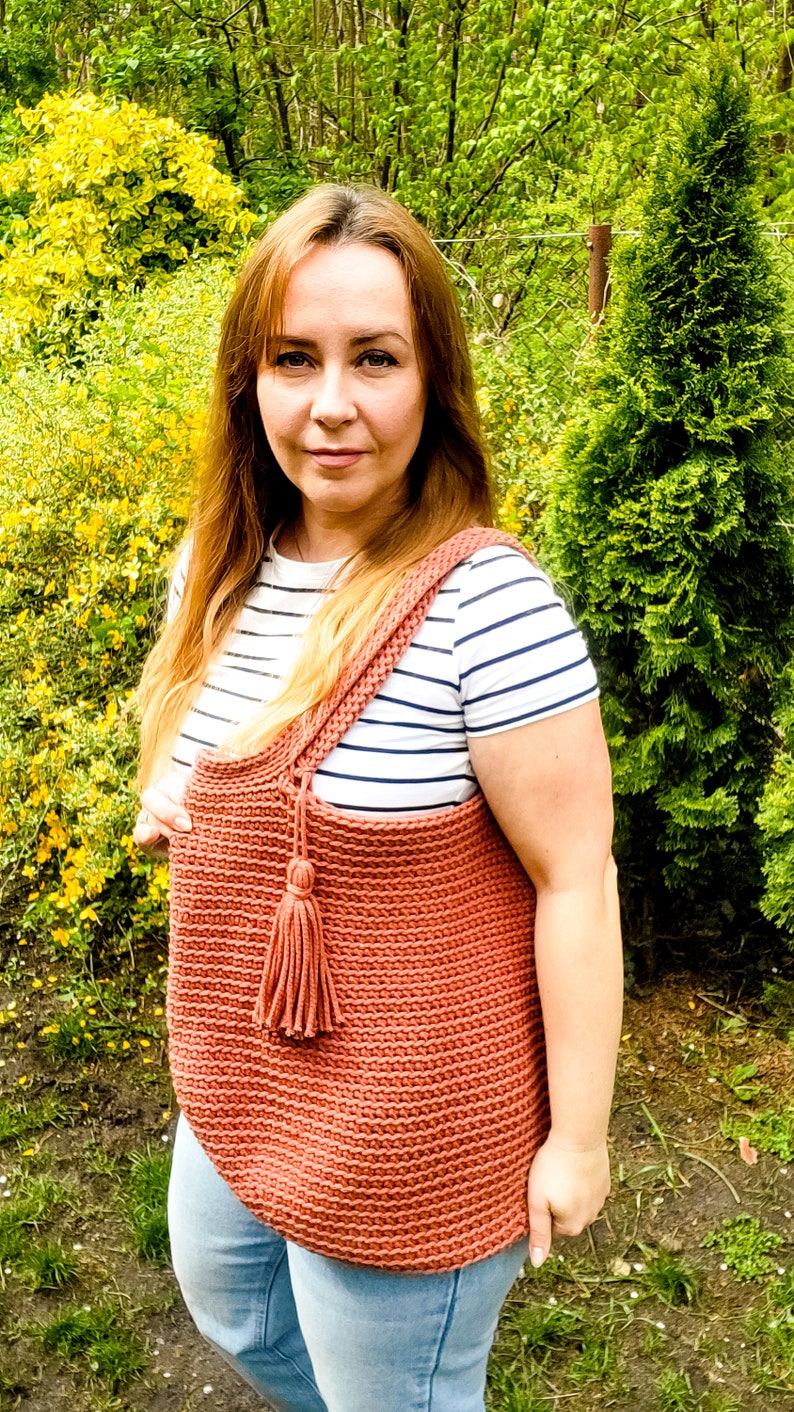 May include: A woman is wearing a striped shirt and blue jeans. She is holding a large, crocheted tote bag with a tassel. The bag is a terracotta color.