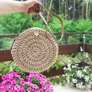 May include: A round, beige crocheted bag with a brown leather strap. The bag has a button closure and is hanging from a hand.