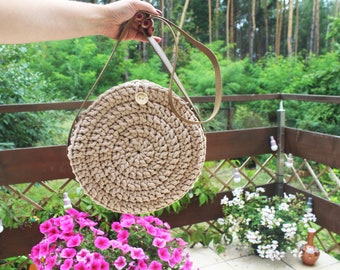 Può includere: Una borsa rotonda in crochet beige con una tracolla in pelle marrone. La borsa ha una chiusura a bottone ed è appesa a una mano.