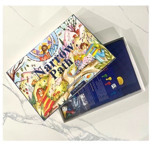 May include: The Narrow Path board game box features a colorful illustration of people walking on a path through a forest. The box is open to reveal the game board and pieces.