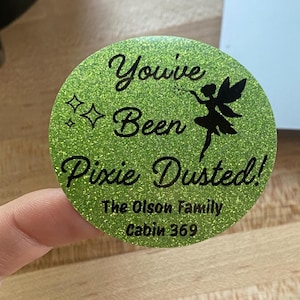 May include: A circular lime green glitter sticker featuring black text that reads You've Been Pixie Dusted! along with a silhouette of a fairy and two small four-pointed stars. Below the main text, it says The Olson Family Cabin 369.