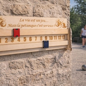 Peut inclure: Tableau de score de pétanque en bois avec des chiffres gravés de 0 à 13. Le tableau comporte un marqueur rouge et un bleu. Le texte "La vie est un jeu... Mais la pétanque c'est serieux !!" est gravé en haut.