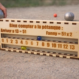 May include: A wooden petanque scoring board with the text "Bien compter a la pétanque" and numbers 0-13. The board has slots for coloured marbles. A person's hand is holding a metal petanque ball. Other petanque balls are in the background.