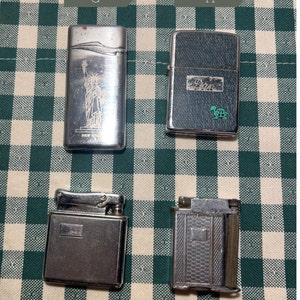 May include: Four silver lighters on a green and white checkered background. The lighters have different designs, including a Statue of Liberty, a turtle, and two with geometric patterns.