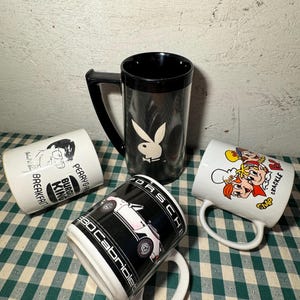May include: Four ceramic mugs with different designs. One mug features a black and white Playboy bunny logo, another has a cartoon image of a burger king, another has a cartoon image of a car, and the last mug has a cartoon image of three people with the text "Snap, Crackle, Pop".