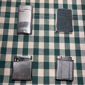 May include: Four silver lighters on a green and white checkered background. The lighters have different designs and textures.