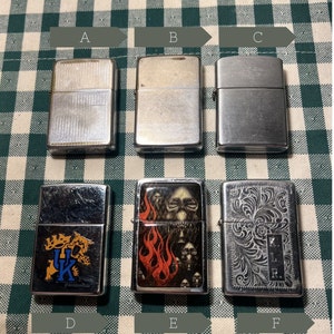 May include: Six silver lighters with different designs. The lighters are arranged in two rows of three. The top row features three plain lighters, while the bottom row features three lighters with designs. The designs include a college logo, a skull with flames, and an intricate floral pattern.
