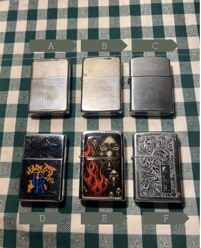 May include: Six silver lighters with different designs. The lighters are arranged in two rows of three. The top row features three plain lighters, while the bottom row features three lighters with designs. The designs include a college logo, a skull with flames, and an intricate floral pattern.