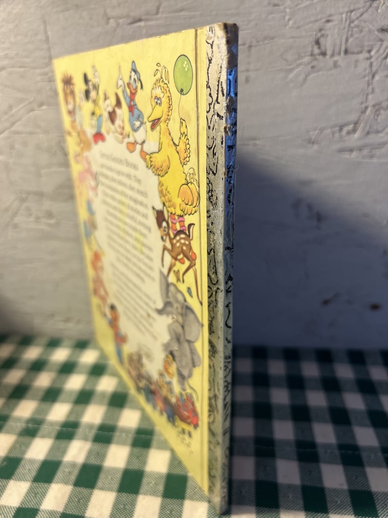 May include: A vintage children's book with a yellow cover featuring a variety of cartoon characters, including Mickey Mouse, Donald Duck, Big Bird, and a deer. The book's title is "The Sesame Street Book of First Words".