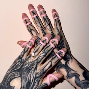 May include: Two hands with detailed black tattoos, wearing pink finger rings. A gold ring with a square diamond is visible. The background is a pale pink.