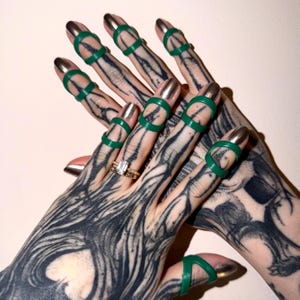 May include: Close-up of hands with silver nail polish and green rings on the fingers. The fingers are adorned with silver nail polish and green rings. The hands are heavily tattooed with black ink.