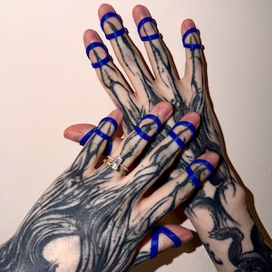 May include: Two hands with detailed black tattoos, wearing blue finger rings. One hand displays a gold ring with a rectangular gemstone. The background is a plain, light-coloured wall.